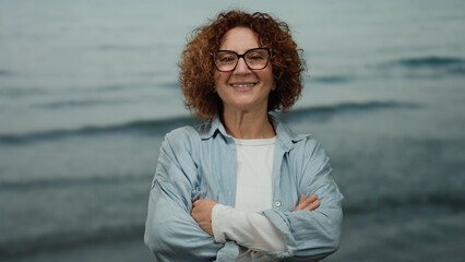 Middle-aged hispanic woman with curly brunette hair and glasses stands smiling with crossed arms...