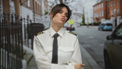Fototapeta premium Young woman pilot in uniform poses playfully on a city street, showcasing confidence and strength in an urban outdoor environment.