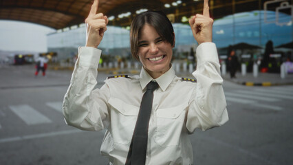 Woman pilot smiling in uniform pointing upwards at airport outdoor showcasing enthusiasm and pride in aviation profession outside terminal building background.