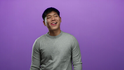 Young chinese man in casual grey sweater making expressive faces against an isolated vibrant purple background wall.