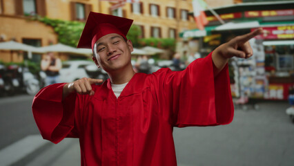 Young man in red graduation gown and mortarboard points and smiles confidently in a bustling city...