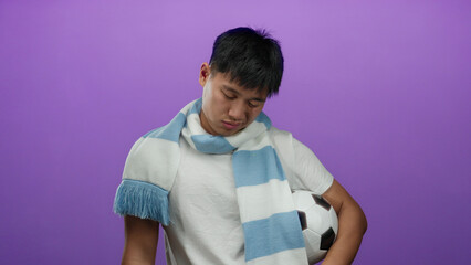 Young chinese man in a white shirt with blue scarf holding soccer ball isolated against purple background, expressing contemplation.