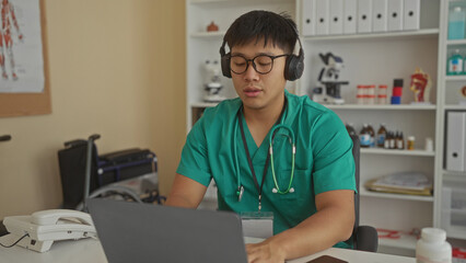 Obraz premium Young man in a clinic wearing scrubs and headphones using a laptop in a workplace setting with medical equipment around.