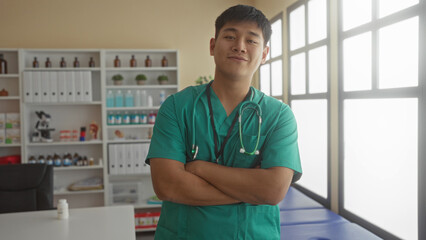 Young doctor man in a clinic room standing with arms crossed, wearing scrubs and stethoscope, displaying confidence in a well-equipped hospital setting with ample natural light.