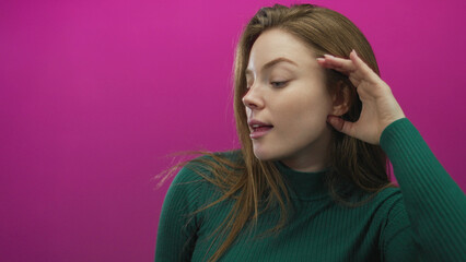 Fototapeta premium Woman in green sweater cups her hand to ear and opens mouth in pink studio with curious tilt of head; surprise attention curiosity intrigue.