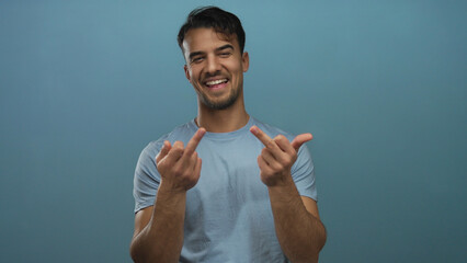 Young hispanic man in a light blue shirt smiling and making a rude gesture against a solid blue...