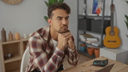 Young man in an office setting analyzing data at a wooden desk with a laptop and guitar in the background conveying focus and contemplation indoors