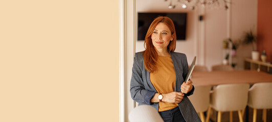 Portrait of pensive female entrepreneur holding laptop and looking away confidently while leaning on wall in office