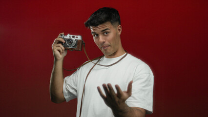 Young man holding vintage camera making come-here gesture over isolated red background, showcasing...