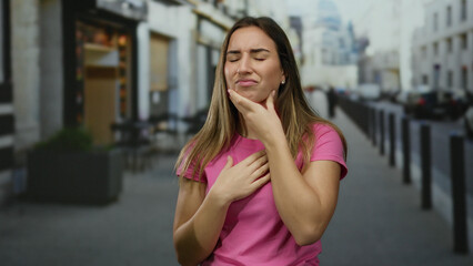 Woman coughing outdoors in urban street wearing pink shirt looking uncomfortable in daylight setting depicting health concern illness.