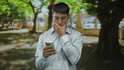 Man in light blue shirt holding smartphone touches chin in green park with sunlight filtering through tree canopy; concern.