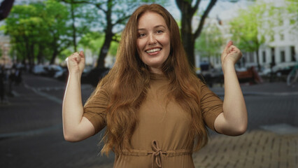 Young caucasian woman in brown dress covers mouth with hands on sunlit urban street; surprise...