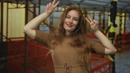 Woman wearing brown dress making peace sign in building under construction, scaffolding behind her;...