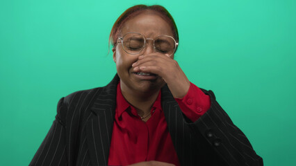 African american woman wearing glasses fights tears and wipes eye with hand in green studio; sadness.