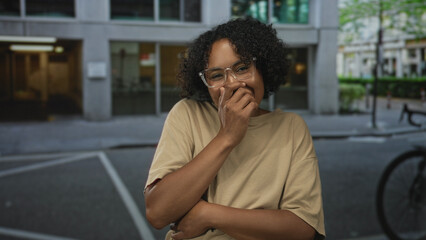 Woman wearing clear glasses and beige shirt covers mouth with hand laughing on busy city street;...