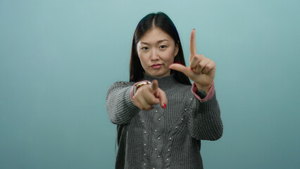 Woman making loser gesture over blue background, emphasizing youthful expression with sweater and...