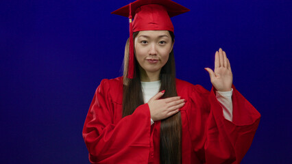 Woman in red graduation gown holding a hand on her chest against a vibrant blue background,...