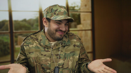 Fototapeta premium Man in camouflage uniform raises palms and shrugs shoulders inside empty building room; uncertainty.