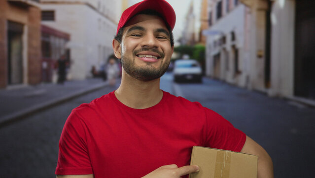 Young hispanic courier man wearing red cap pointing finger to package on busy city street; service reliability.