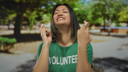 Naklejka premium Young chinese woman volunteer in green shirt crossing fingers outdoors in a park setting, conveying hope and optimism under bright sunlight with blurred trees in the background.