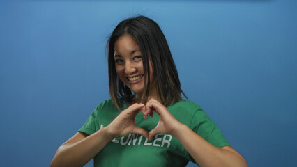 Young woman smiling while making heart gesture wearing volunteer t-shirt against vibrant blue...