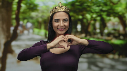 Woman wearing crown in park making heart gesture with hands, showcasing joy and self-love, surrounded by lush trees, embodying elegance and playfulness in a serene outdoor setting.