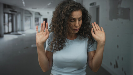 Young hispanic woman holds up both hands with palms facing outward inside an empty building;...