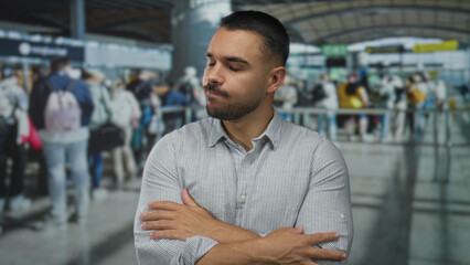 Hispanic man in striped buttonup shirt closes his eyes and crosses his arms amid travelers at...