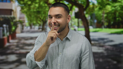 Man bringing finger to lips for silence on city street under leafy trees in bright sunlight;...