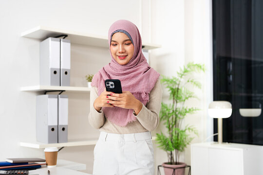 Smiling Muslim business woman in hijab using mobile phone  standing in office. Busy young businesswoman manager, female executive leader holding mobile phone at work.