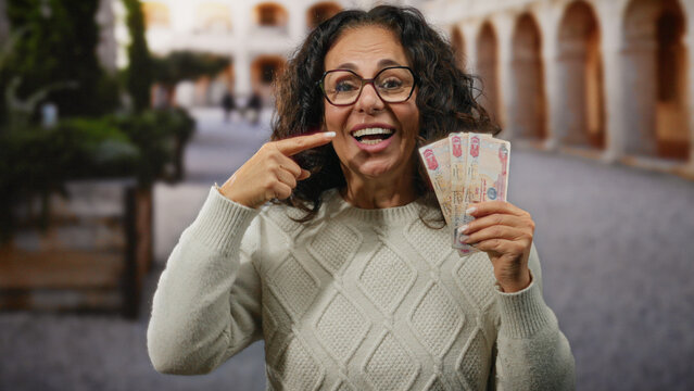 Middle-aged woman in uae holding dirham banknotes and pointing to her teeth, standing outdoors in a city street.