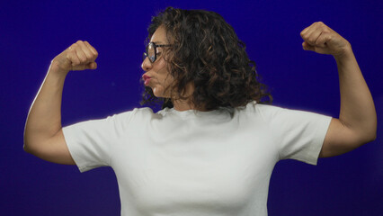 Hispanic middle-aged woman confidently flexing her muscles against an isolated blue background...