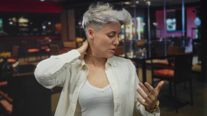 Woman with short gray hair holds hand to forehead with sweat sheen under warm overhead lights in building; discomfort.