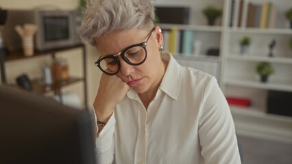 Woman with short silver hair and round glasses resting head on hand at office desk with computer...