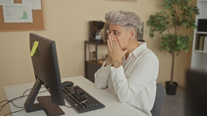 Woman removes glasses at office desk by computer monitor against corkboard in modern office...
