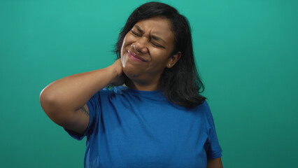 Woman holds neck with one hand and winces with closed eyes, wearing blue shirt in teal studio;...