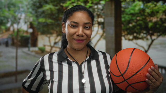 Woman referee smiles and holds basketball with whistle on green street lined with foliage; confidence. - Powered by Adobe
