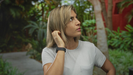 Blonde woman resting hand on chin wearing smartwatch and t shirt in green park under sunlight;...
