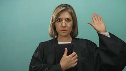 Young blonde woman holding a black book and raising her hand in a teal studio setting; pledge...