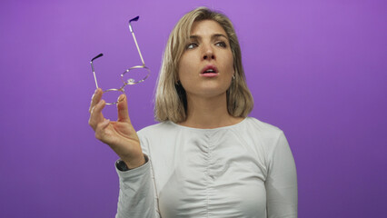 Woman blonde young removes round gold glasses with hand while squinting in purple studio wearing...