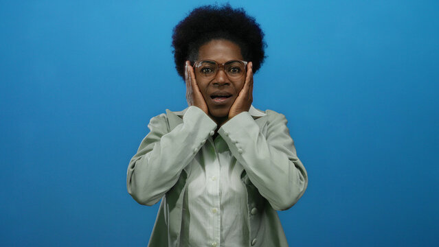 Woman in glasses shows surprise over isolated blue background, communicating emotion, focus, expression, and diversity through visual language and professional attire.