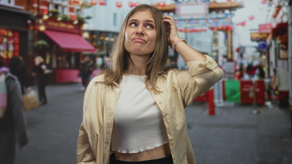 Young blonde woman with hand in hair and raised eyebrow on a bustling city street decorated with...