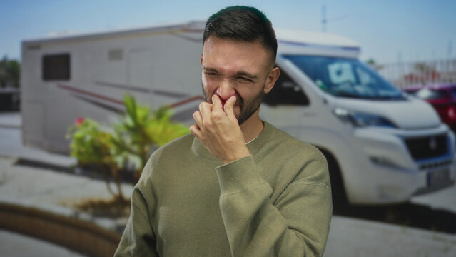 Young hispanic man outdoors pinching nose on street due to bad smell near rv urban area sunny day showing discomfort.