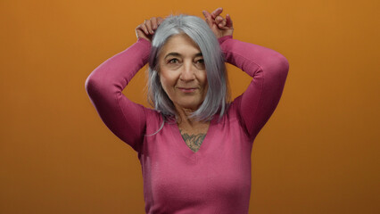 Senior woman with grey hair playfully gestures with hands on an orange background, wearing a pink...