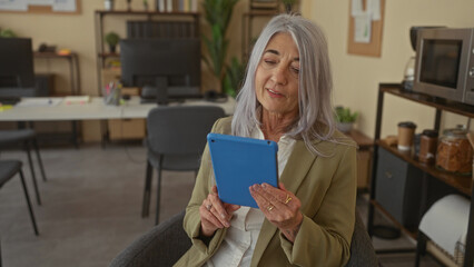 Senior woman with grey hair holding tablet in modern office setting, surrounded by contemporary...