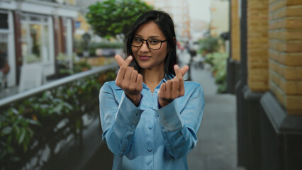 Woman in blue shirt making heart gestures outdoors on a busy street with glasses and dark hair...