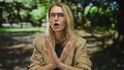 Woman with blonde hair and glasses in a park makes an x gesture with her arms in a green outdoor...
