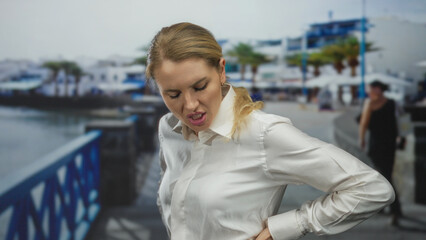 Woman standing on seaside promenade in discomfort wearing white shirt with hand on back against...
