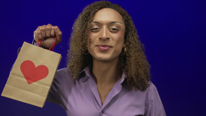 Woman holds shopping bag with heart graphic, smiles broadly and raises fist in studio; excitement...