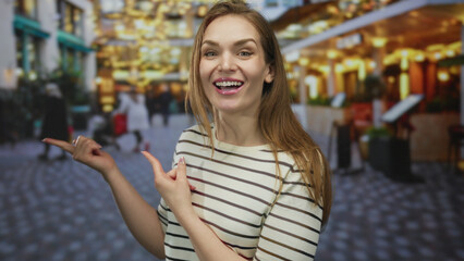 Young woman in striped top smiling and pointing finger to side on busy street under glowing cafe...